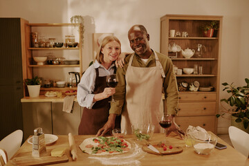 Senior Caucasian woman and senior Black man standing together in kitchen preparing homemade pizza, smiling and looking at camera, holding wine glass, showing romantic connection