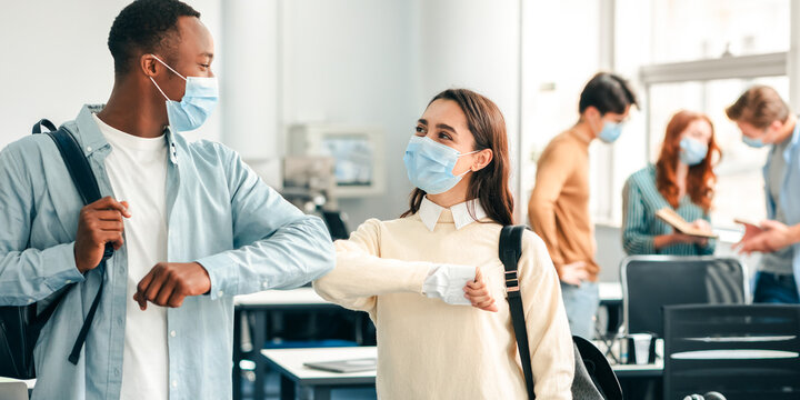 Elbow Bump. Smiling multicultural students wearing protective face masks and backpacks greeting each other. Woman and african american guy avoid touching, studying in university, standing at classroom