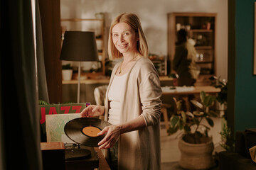 Caucasian senior woman smiling while holding vinyl record near turntable, standing in cozy living...