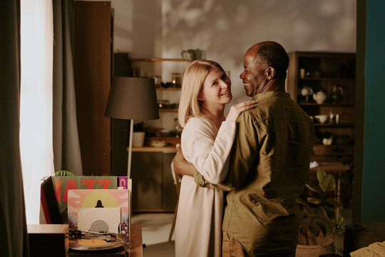 Senior Caucasian woman and senior Black man embracing while dancing together in living room, smiling and making eye contact, expressing romantic connection and affection - Powered by Adobe
