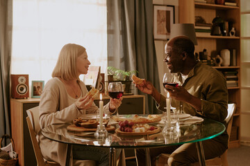 Senior Caucasian woman and senior Black man sitting at glass table sharing meal and wine, smiling at each other, holding food in hands, enjoying romantic moment together indoors