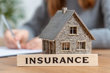 A detailed miniature house sits on a wooden table with the word insurance prominently displayed in front.