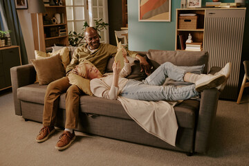 Senior Caucasian woman lying on sofa with head on lap of senior Black man smiling, both relaxing together while holding digital tablet, showing romantic moment in home setting