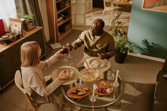 Senior Caucasian woman and senior Black man sitting at dining table sharing romantic dinner, smiling and clinking wine glasses, enjoying meal together in cozy home setting - Powered by Adobe