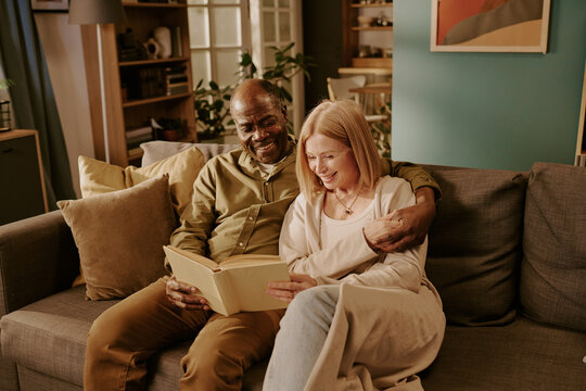 Senior Black man and senior Caucasian woman sitting on sofa smiling and reading book together, man embracing woman, couple enjoying romantic moment in cozy living room setting