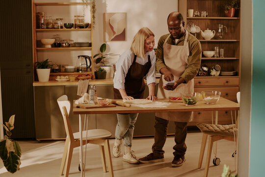 Senior Caucasian woman and senior Black man preparing dough together in kitchen, smiling and interacting while baking, wooden table with ingredients visible, couple enjoying shared activity