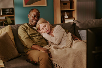 Senior Black man and senior Caucasian woman sitting on sofa, smiling and relaxing together, woman resting head on man's shoulder, couple watching television, showing romantic connection