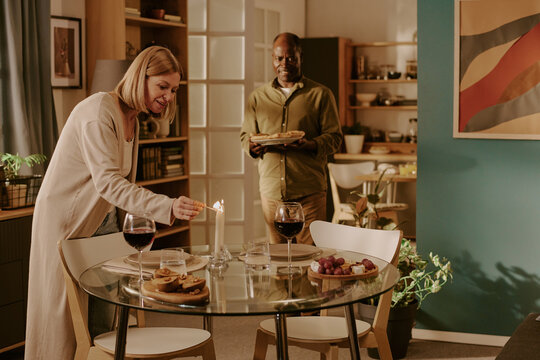 Senior Caucasian woman lighting candle on dining table while senior Black man holding plates approaching, both preparing romantic dinner together in cozy home setting - Powered by Adobe