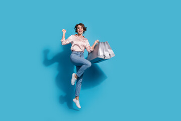 Joyful young woman in pink blouse jumping with shopping bags against blue background
