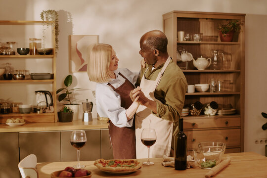 Senior Caucasian woman and senior Black man dancing together in kitchen, smiling and holding each other, wine glasses and pizza on table, enjoying romantic moment at home - Powered by Adobe