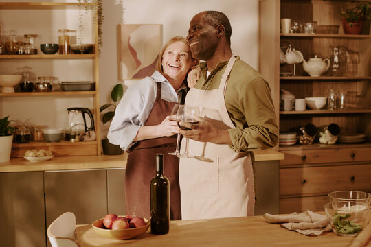 Senior Caucasian woman and senior Black man standing together in kitchen smiling and embracing while holding wine glasses, showing romantic connection and enjoying each other's company - Powered by Adobe