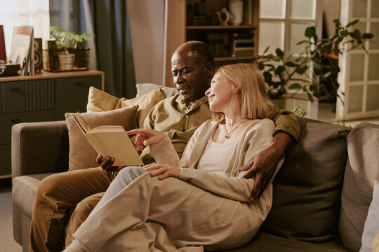 Senior Black man and senior Caucasian woman sitting together on sofa reading book, smiling and embracing, showing romantic connection and enjoying shared moment in cozy living room - Powered by Adobe