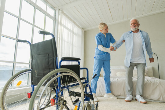 Rehabilitation healthcare. Physiotherapist nurse helps old man patient in wheelchair stand up walking. Man in chair for people with disability doing few steps with nurse helping. Medical caregiver