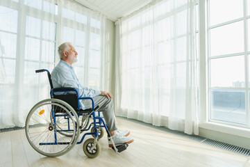 Sad senior man sitting in wheelchair looking out window in retirement home hospital. Old paralyzed man in chair for people with disability feel depressed. Lonely elderly sick man sitting on wheelchair