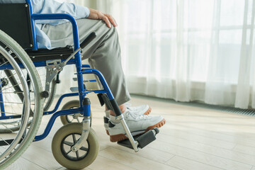 Senior man sitting in wheelchair near window in retirement hospital, legs feet close up. Old paralyzed man in chair for people with disability at home. Lonely elderly sick man sitting on wheelchair