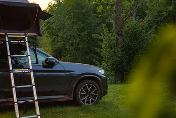 A dark colored SUV with a foldable rooftop tent and ladder parked on grass in a forest. Dirt on the vehicle suggests recent off road travel.