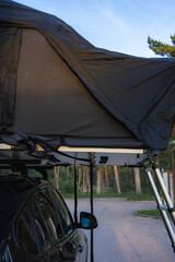 A modern SUV with a fully extended rooftop tent parked on a paved surface in a forest. Natural light illuminates the trees and camping setup.