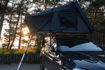 A modern SUV with a foldable rooftop tent and telescopic ladder is parked on a paved surface in a forest. Tall trees and sunlight create a serene setting.
