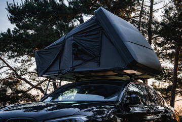 A modern SUV with a dark colored rooftop tent set up in a forest. Tall trees surround the vehicle as soft light filters through the branches.