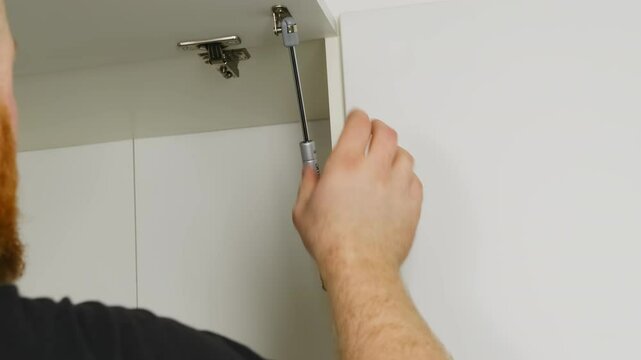 Master installs furniture shock absorber during work in flat. Specialist provides smooth opening and closing of cabinet door analyzing mechanism condition