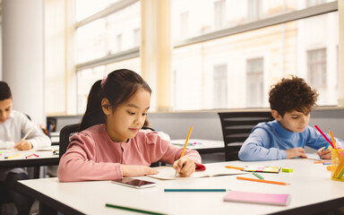 Learning Process Concept. Portrait of Small Schoolchildren Smiling, Sitting At Desk Taking Notes And Writing in Exercise Copybook. Junior Classroom with Diverse Group of Children Learning New Stuff