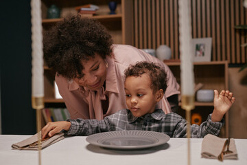 Smiling Black woman talking to child in dining room during festive dinner while child interacting with table setting