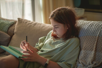 Concentrating on her digital art, a preteen girl in casual clothes draws on a tablet with a stylus, enjoying leisure time while relaxing on a cozy sofa at home