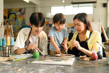 A toddler joins in painting during an art class, guided by enthusiastic parents and teacher in a...