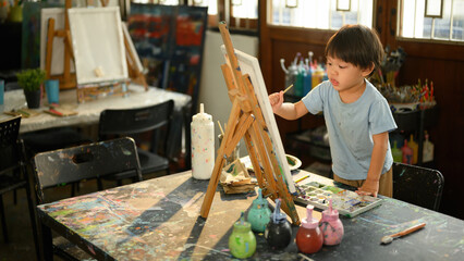 A preschool boy focused on painting at an easel in a bright and colorful art classroom