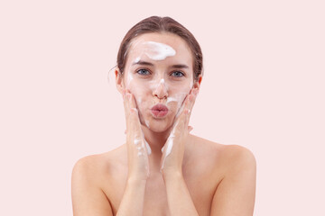 Young woman washing her face with foaming cleanser, hands covered in foam touching her skin. She...