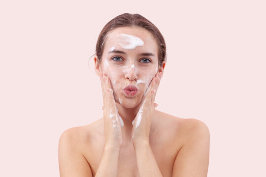 Young woman washing her face with foaming cleanser, hands covered in foam touching her skin. She blows a kiss toward the camera. Skincare routine for clean, healthy face. Isolated on pink background