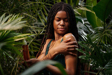 Black woman applying skincare product on her shoulder while standing amidst lush greenery. Relaxed...