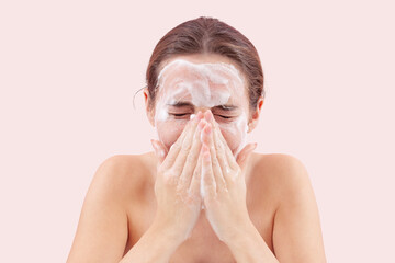 Close up of a young woman washing her face with hands covered in foam touching her skin. Deep cleansing skincare routine for clean, healthy skin. Isolated on pink background, acne care concept