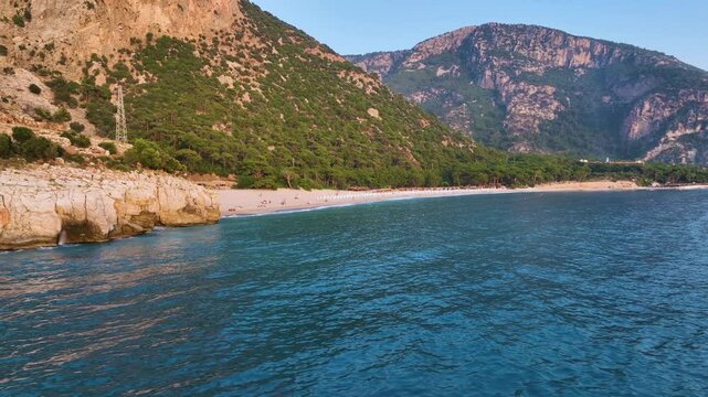 Calm turquoise water gently laps the sandy shore of phaselis beach, nestled against a backdrop of lush green mountains, as tourists relax and enjoy the serene sunset