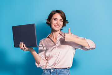 Smiling young woman holding a laptop giving thumbs up with blue background, expressing confidence and success