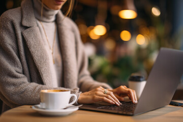 Fototapeta premium Businesswoman in smart casual outfit working remotely from a stylish café, typing on laptop, cup of coffee beside her, background softly blurred with warm tones freelance