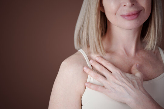 Smiling Caucasian woman with shoulder-length blonde hair wearing beige tank top and touching collarbone with her hand, highlighting emotion of calmness