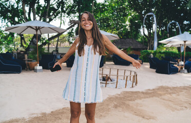 Joyful young woman spreading arms and laughing while standing on tropical resort sand with...