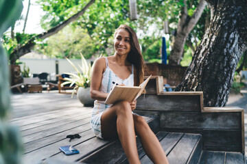Confident young female freelancer reflects in notebook, bridging digital tools and analog thought...