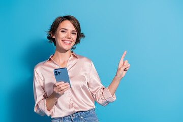 Smiling young woman in stylish satin blouse pointing at copy space against a blue background,...