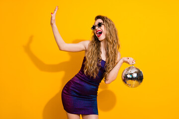 Happy young woman in stylish dress holding a disco ball on a vibrant yellow background, celebrating...