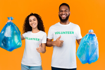 Couple Of Multiethnic Volunteers Holding Blue Plastic Garbage Bags Picking Litter Gesturing Like Standing Over Yellow Studio Background. Volunteering Work, Let's Keep Planet Environment Clean Concept