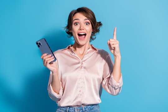 Young woman in pink blouse excitedly holding a smartphone with a surprised smile, pointing up against a vibrant blue background