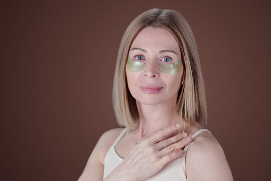 Portrait of woman wearing under-eye patches for skincare on brown background, showing relaxed expression. Clear skin visible, hand touching neck, route to natural beauty highlighted - Powered by Adobe