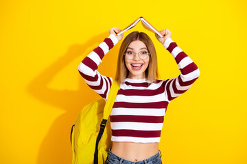 Smiling young woman wearing striped pullover holding book over head, standing against bright yellow...
