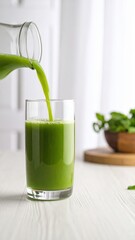 pouring fresh green juice into glass on white wooden table with bowl of greens behind it