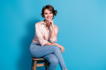Young stylish woman in pink shirt and jeans smiling against blue background, sitting confidently on...