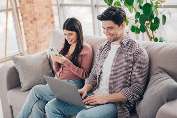 Obraz premium Young couple relaxing at home together, using a smartphone and laptop on a comfortable sofa in a bright modern living room interior.