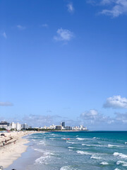 Fototapeta premium A bright, sunny view of Miami Beach with turquoise ocean waves, white sandy shore, and a backdrop of hotels and high-rise buildings along the coastline. 