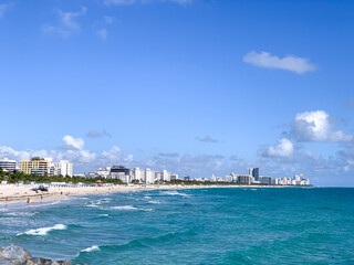 Fototapeta premium A bright, sunny view of Miami Beach with turquoise ocean waves, white sandy shore, and a backdrop of hotels and high-rise buildings along the coastline. 
